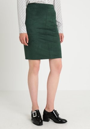 Jupe crayon en daim vert avec des coutures verticales ; associée à des chaussures en cuir verni noir avec un détail de boucle carrée.