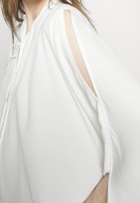 Blusa bianca con una texture morbida, caratterizzata da aperture alle spalle e una chiusura a fiocco al collo. Design a vestibilità ampia.