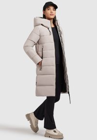 Manteau long en duvet gris clair avec capuche, fermeture éclair à l'avant, poches latérales et design matelassé, associé à un pantalon noir et des baskets crèmes épaisses.