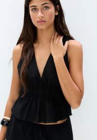 Top senza maniche nero con scollo a V profondo, vita arricciata e orlo sfilacciato. Presenta un tessuto leggero con una silhouette rilassata.