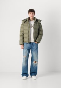 Olivengrønn boblejakke med hette, med kviltet design. Båret over en lys grå sweatshirt og kombinert med slitte blå jeans.
