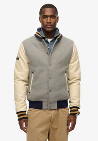 Chaqueta bomber de mezcla de lana gris con mangas de cuero crema, puños de canalé azul marino y cuello a rayas. Bolsillos frontales y cierre de botones a presión.