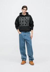 Fekete kapucnis pulóver nagy fehér betűkkel az "OBEY" felirattal, laza kék farmerral és világosbarna lábbelivel párosítva. Egyszerű háttér, a modell zsebre tett kézzel áll.
