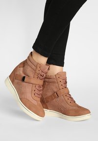 Hnědé wedge tenisky se šňůrkami a bočními popruhy, nošené s černými kalhotami, zobrazené na zkřížených nohách na bílém pozadí.