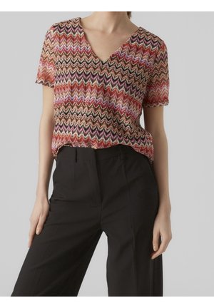 Femme portant un haut à manches courtes en V à motif zigzag multicolore et un pantalon noir taille haute, les mains reposant sur les côtés.