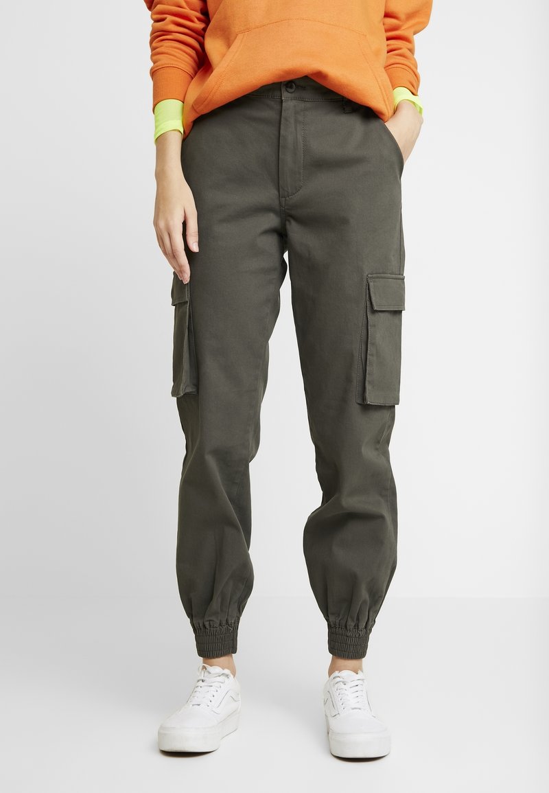Pantalon cargo vert olive avec bas élastiques, doté de poches latérales et d'une coupe décontractée, porté avec des baskets blanches et un sweat à capuche orange.