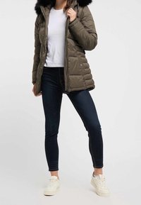 Doudoune vert olive avec capuche en fourrure noire, fermeture éclair et design matelassé. Associée à un jean foncé et des baskets blanches.