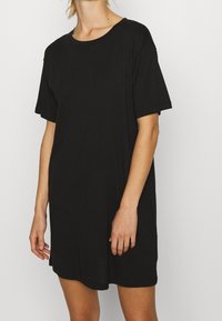 Femme portant une robe T-shirt noire à manches courtes ample, se tenant devant un fond blanc uni.