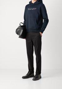 Námořnická mikina s přední kapsou a logem, černé kalhoty a černé boty. Model drží texturovanou černou tašku. Jednoduchý a ležérní outfit.