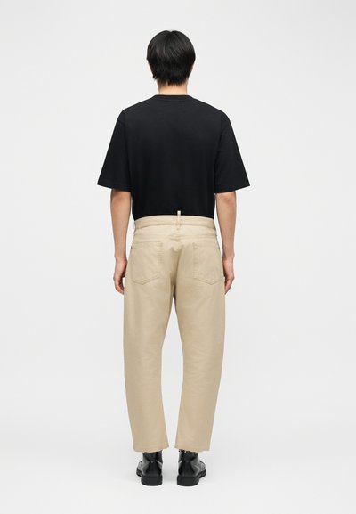 Personne debout, vue de dos, portant une chemise noire à manches courtes, un pantalon beige et des bottes noires, sur un fond clair uni.