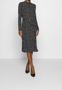 Svart klänning med lång ärm som har ett vitt geometriskt mönster, insnörad midja och utsvängd nederkant, tillsammans med spetsiga svarta klackar.