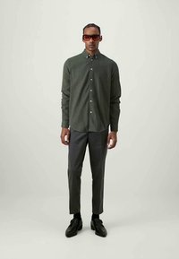 Camisa verde de botão com um acabamento texturizado, combinada com calças cinza-escuras ajustadas e sapatos pretos. Óculos de sol grandes em tom castanho incluídos.