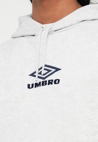 Primo piano di una persona che indossa un felpa con cappuccio grigio chiaro, con il logo Umbro blu scuro e i lacci sul petto.
