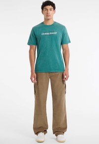 Teal bombažna majica s kratkimi rokavi in okroglim izrezom, na kateri je bel napis "GUESS JEANS". V kombinaciji s svetlo rjavimi cargo hlačami in belimi športnimi copati.