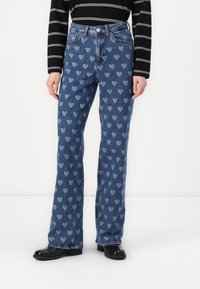 Svetlo modri kavbojke z ponovljenim vzorcem srca. Oblikovanje s širokimi hlačnicami, pet žepov in gumbom za zapiranje. Črne gležnjarje.