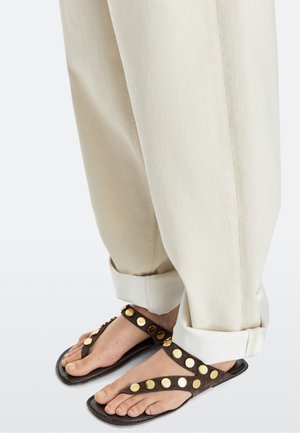 Pieds portant des sandales marron foncé avec de gros clous dorés, associés à un pantalon beige clair retroussé, sur un fond clair uni.