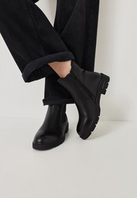 Černé kožené kotníkové boty se zipem na boku, s robustní podrážkou a texturovaným povrchem, spárované s ohrnutými černými džínovými kalhotami.