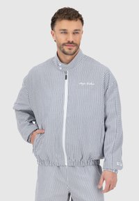 Veste rayée bleu et blanc, dotée d'un col montant, d'une fermeture éclair et de poignets élastiques. Le matériau semble texturé avec une coupe décontractée.