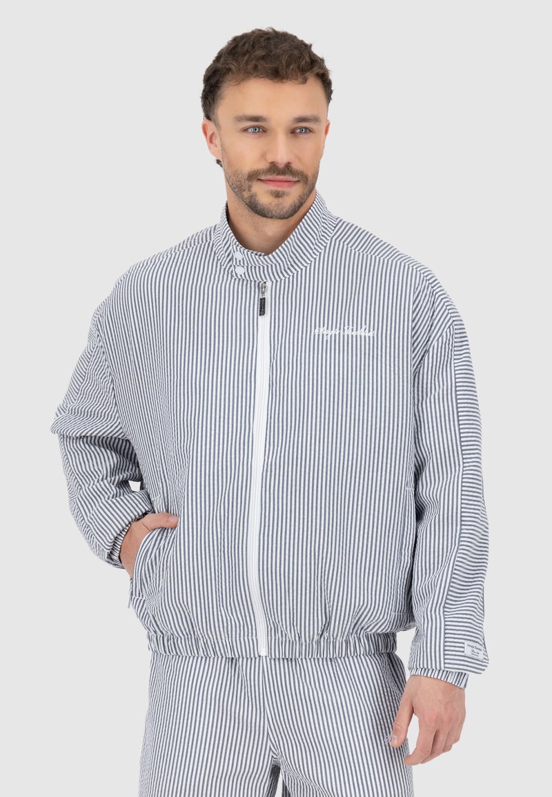 Veste rayée bleu et blanc, dotée d'un col montant, d'une fermeture éclair et de poignets élastiques. Le matériau semble texturé avec une coupe décontractée.