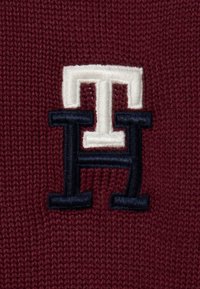Burgundowy dzianinowy materiał z wyraźnym haftowanym logotypem w bieli i granacie, przedstawiającym litery 'H' i 'TH' w warstwowym wzorze.