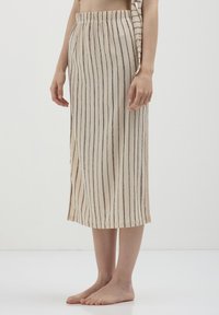 Femme portant une jupe beige avec des rayures verticales noires, debout pieds nus sur un sol blanc contre un fond blanc uni.