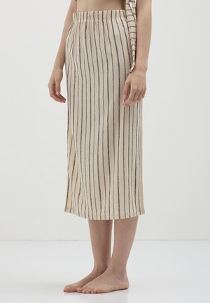 Donna che indossa una gonna beige con strisce verticali nere, in piedi a piedi nudi su un pavimento bianco contro uno sfondo bianco semplice.
