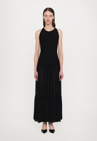 Robe noire avec un corsage ajusté à bretelles et une jupe maxi plissée transparente, présentant des volants superposés au niveau de l'ourlet et des talons aiguilles noirs.