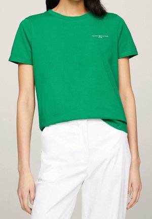 Kobieta ubrana w zielony krótkim rękawem t-shirt Tommy Hilfiger i białe spodnie na jednolitym tle.