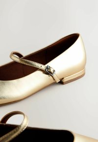 Zapatos planos metálicos dorados con punta redonda y una sola correa ajustable. Presentan un pequeño tacón cuadrado y una textura suave.