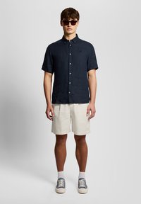 Νεαρός άνδρας που φορά σκούρο μπλε πουκάμισο με κοντά μανίκια και κουμπιά, μπεζ σορτς, λευκές κάλτσες, μπλε και λευκά αθλητικά παπούτσια και σκούρα γυαλιά ηλίου, όρθιος.