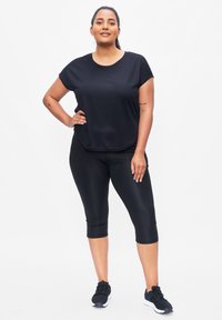 Zwart sporttop met korte mouwen en een ronde hals, gecombineerd met zwarte capri leggings. Beide zijn gemaakt van een gladde, rekbare stof. Zwarte sportschoenen.