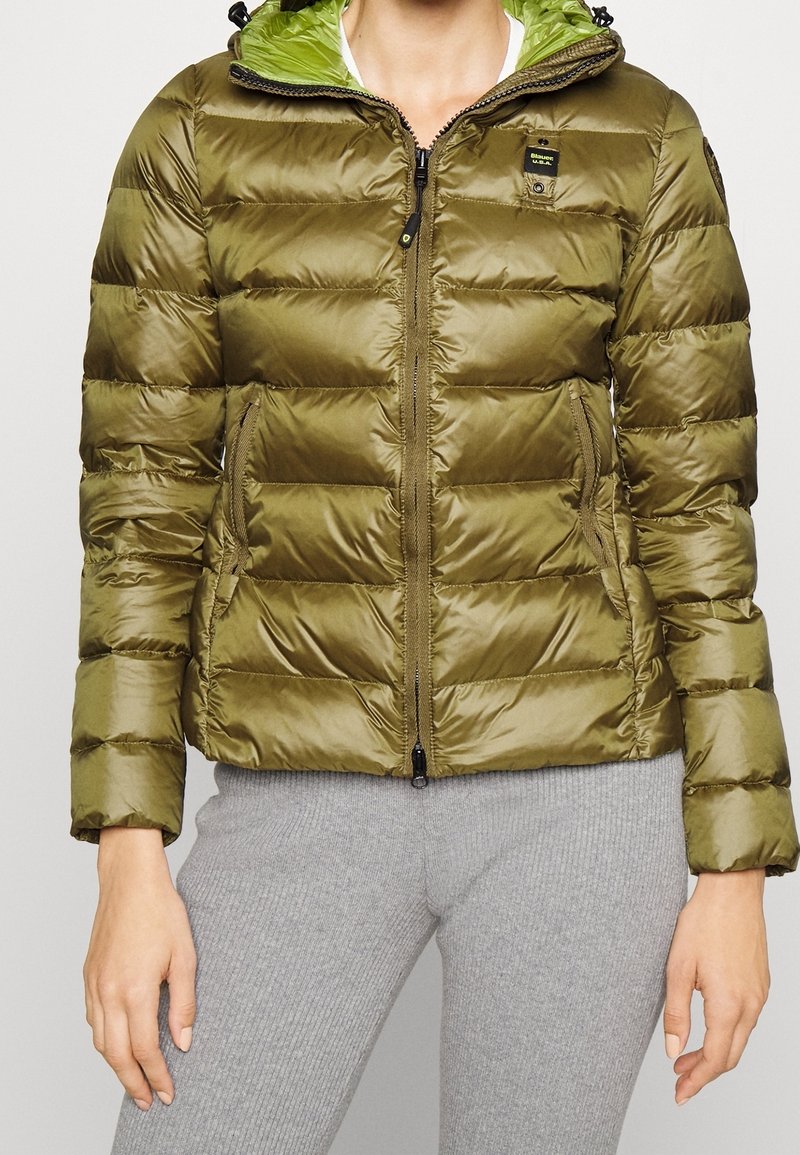 Mujer con chaqueta acolchada tipo puffer color verde oliva con cremallera frontal, bolsillos laterales y capucha verde, combinada con pantalones grises acanalados.