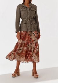 Khakifärgad arbetsjacka med fyra fickor framtill och dragsko i midjan, kombinerad med en midjelång blommönstrad kjol i jordnära toner. Beige blockklackar.