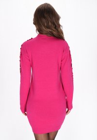 Donna con lunghi capelli castani che indossa un vestito a maniche lunghe di maglia rosa brillante con dettagli di paillettes sulle maniche, girata di spalle su uno sfondo semplice.