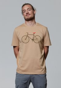 Beige t-shirt van zacht materiaal met een grafisch ontwerp van een fiets in bruin met groene en oranje accenten. Korte mouwen, relaxte pasvorm.