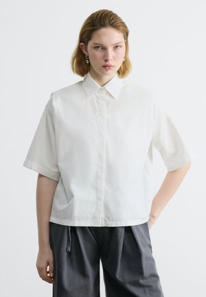 Young person with short blonde hair wears white short-sleeve collared shirt and dark gray loose pants, standing with one hand on hip.