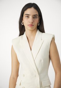 Blazer sans manches croisé en tissu crème avec une finition texturée, doté d'accents aux épaules et de poches latérales.