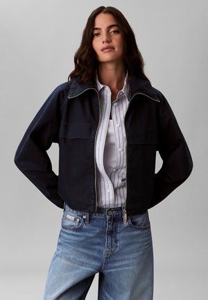 Kobieta o falowanych ciemnych włosach, ubrana w czarną kurtkę na zamek i biało-paskowaną koszulę na guziki oraz niebieskie dżinsy, stojąca z rękami za plecami.