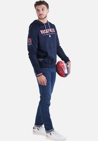 Haut à capuche marine avec des accents rouges et le numéro 8, associé à un jean bleu et des baskets blanches. Tient un ballon de rugby aux couleurs variées et avec des marques.