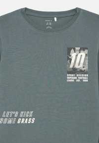 Camiseta de algodão verde com decote redondo, estampas gráficas com o número "10" e texto a dizer "VAMOS CHUTAR ALGUMA GRAMA."