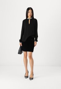Blouse noire avec un décolleté en forme de keyhole et des manches longues, associée à une jupe noire ajustée en quilté. Accentuée d'un collier argenté et d'un sac à main noir.