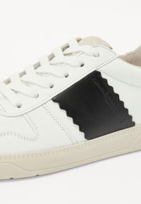 Sneaker bianco con un accento in pelle nera texturizzata e motivo a zigzag, lacci piatti e suola in gomma con dettagli angolari.