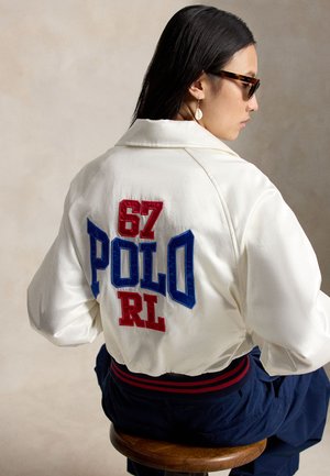 Nainen valkoisessa takissa, jossa takana punaisilla ja sinisillä kirjaimilla "67 POLO RL", istuu puisella jakkaralla, aurinkolasit päässä ja roikkuvat korvakorut.