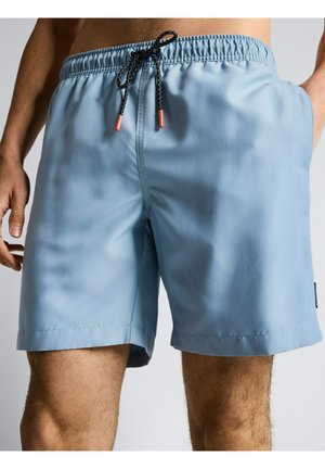 Hellblaue Herren-Badeshorts mit schwarzem und weißem Kordelzug und orangen Spitzen, getragen von einem stehenden Mann mit entspannten Händen.