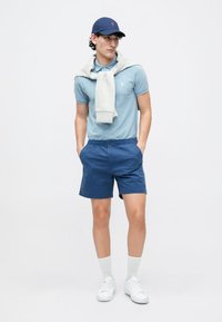 Light blue polo shirt, navy shorts, grey sweater draped around shoulders, navy cap, white sneakers, and socks. Simple, casual design.