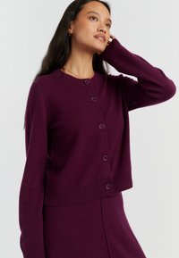 Cardigan in lana viola con scollatura a girocollo, chiusura con bottoni, orli a coste e maniche lunghe. Il tessuto sembra morbido e con una superficie texturizzata.