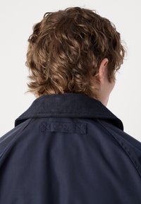 Personne aux cheveux bruns courts et bouclés portant une veste bleu marine avec un col en velours côtelé relevé, vue de dos.
