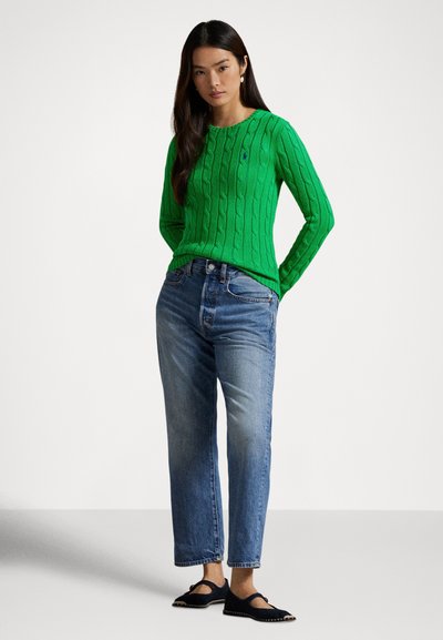 Grønn kabelstrikket genser med lange ermer, brukt sammen med lyseblå jeans med rette ben og svarte flate sko. Enkel rund hals.