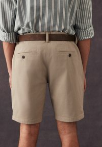 Pantalones cortos beige con una textura tejida, dos bolsillos traseros con cierres de botón y un cinturón marrón. dobladillo visible y corte entallado.