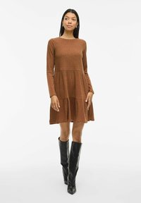 Braunes, geripptes Langarmkleid mit weit schwingendem Saum und gestuftem Design, kombiniert mit schwarzen kniehohen Stiefeln. Glatte Textur und taillierter Oberkörper.